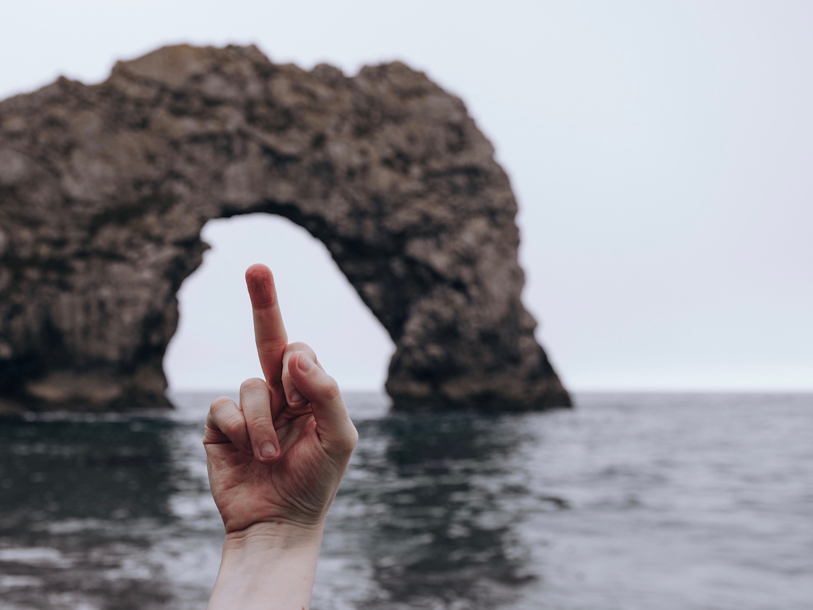 Palm-forward middle finger raised in front of Durdle Door by the sea